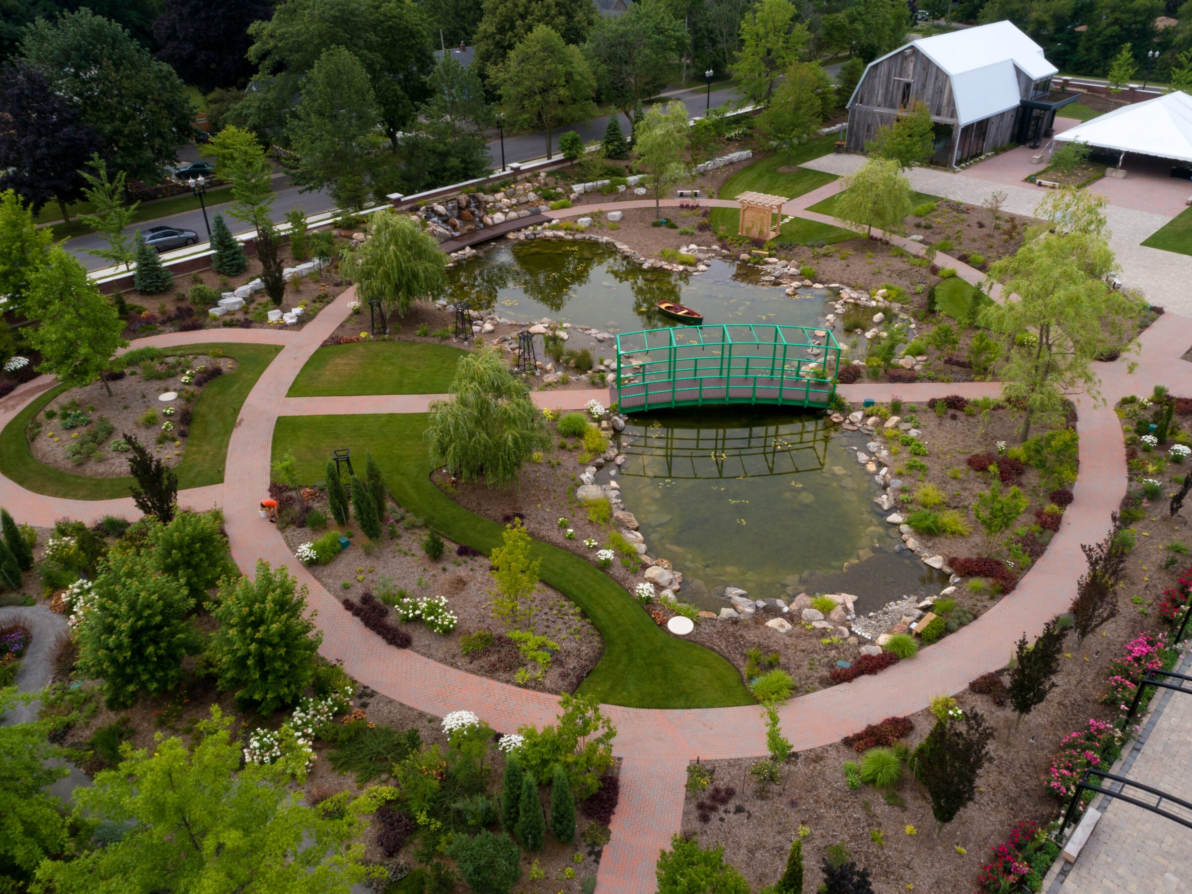 Aerial view of a landscaped garden with winding pathways, pond, green bridge, lush foliage, and a rustic barn surrounded by trees.
