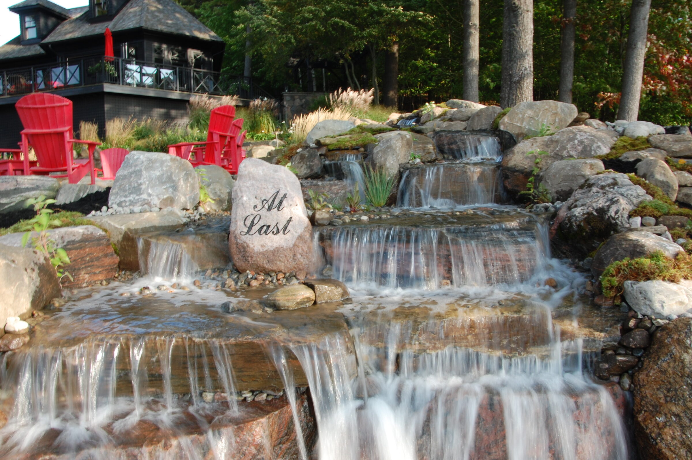 Tranquil waterfall flows over rocks near red chairs and stone with “At Last” engraved, surrounded by greenery and a dark house in the background.