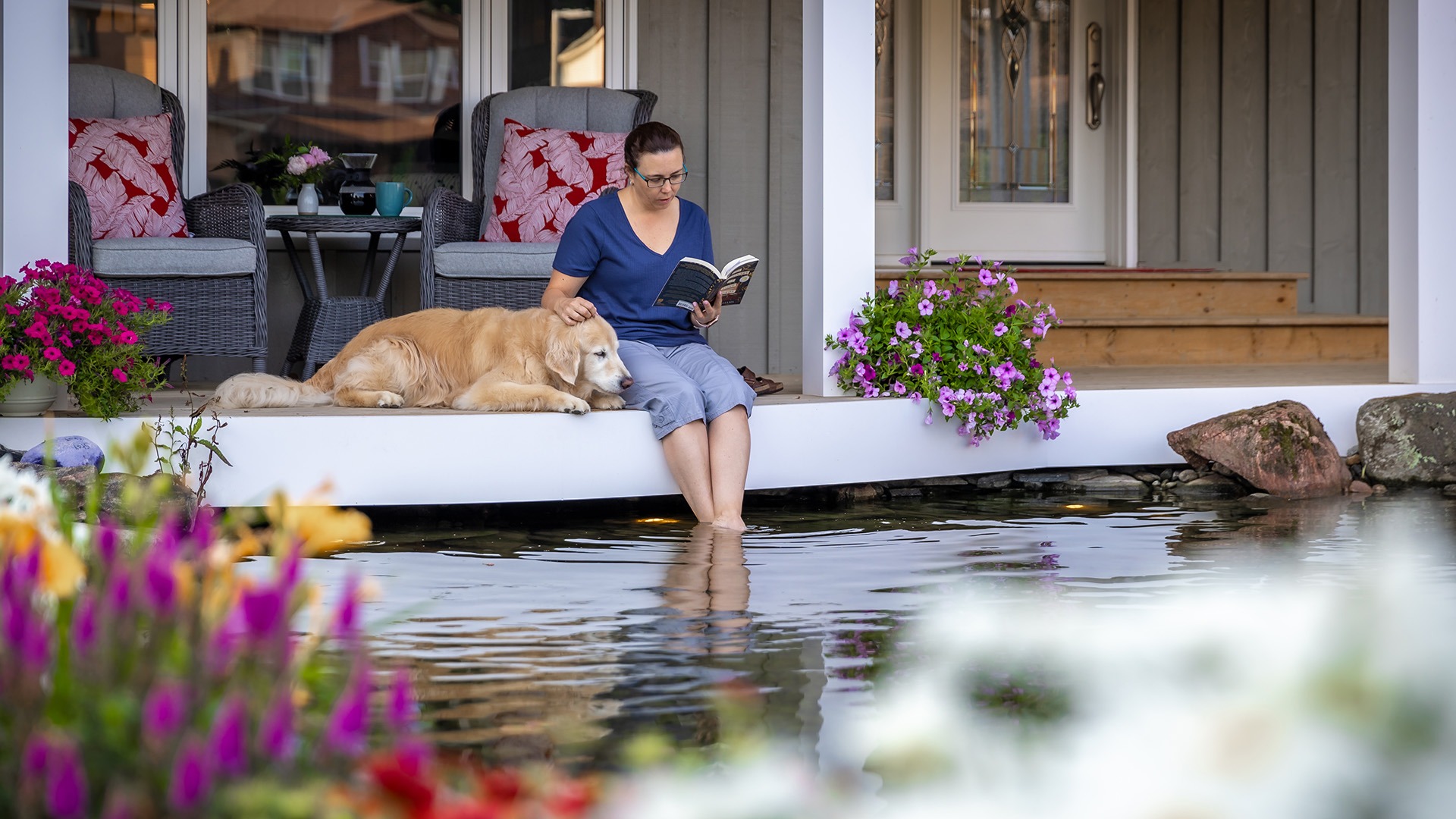 A person reads by a pond, petting a golden retriever on a porch adorned with vibrant flowers and comfortable seating.
