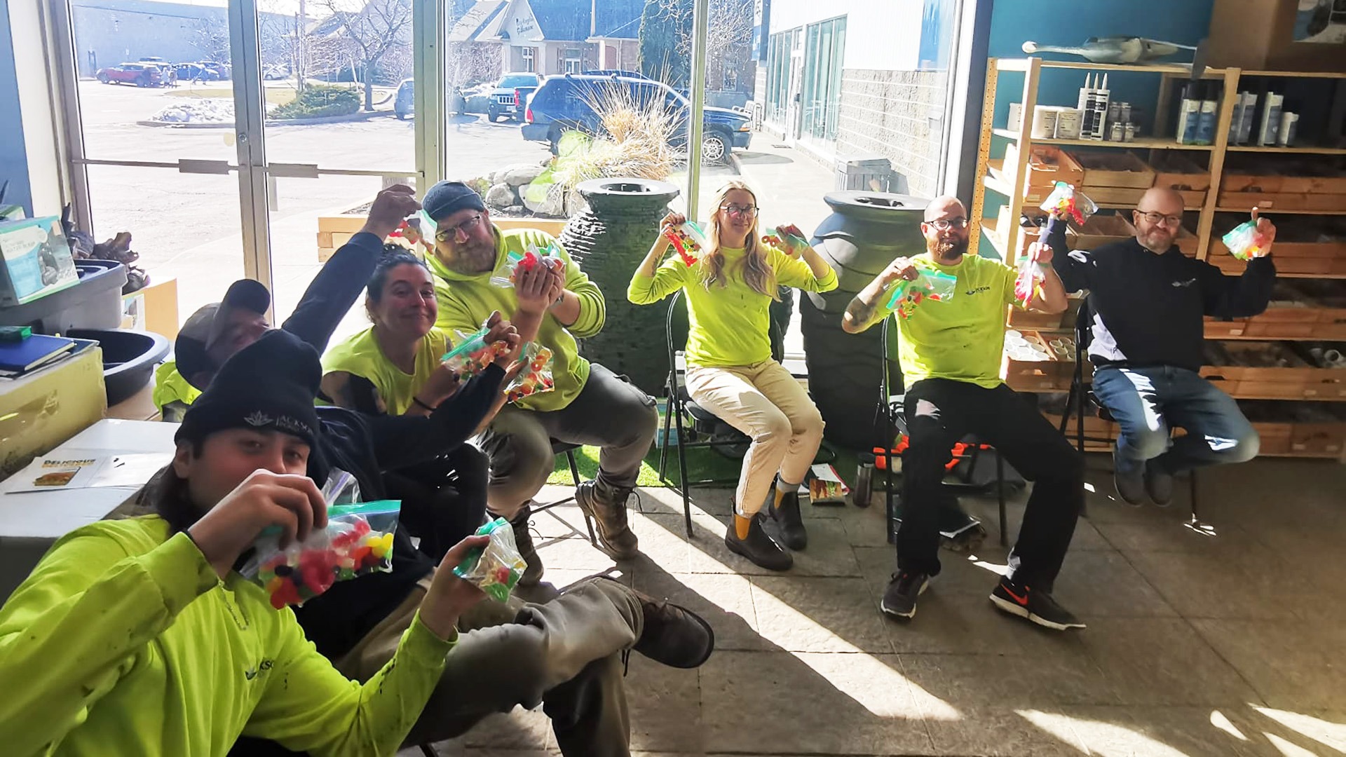 A group of people in bright shirts sit indoors, each holding a colorful bag of candy, smiling in a sunlit room.