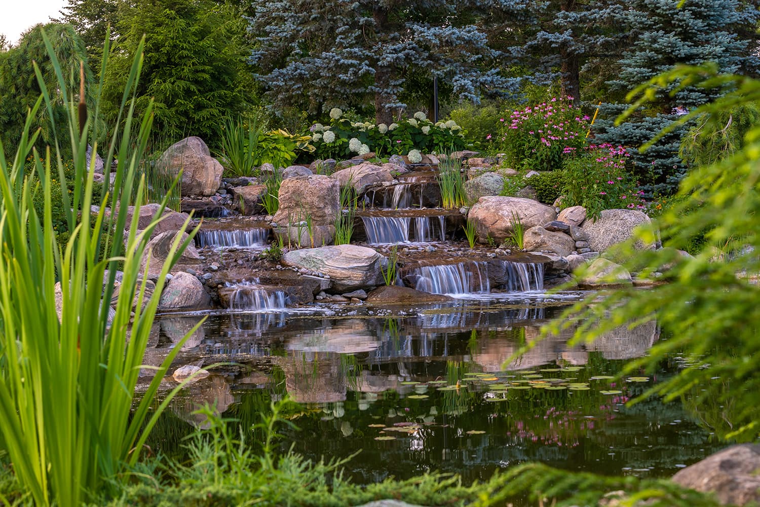A serene garden features cascading waterfalls over rocks into a reflective pond, surrounded by lush green plants and vibrant flowers.