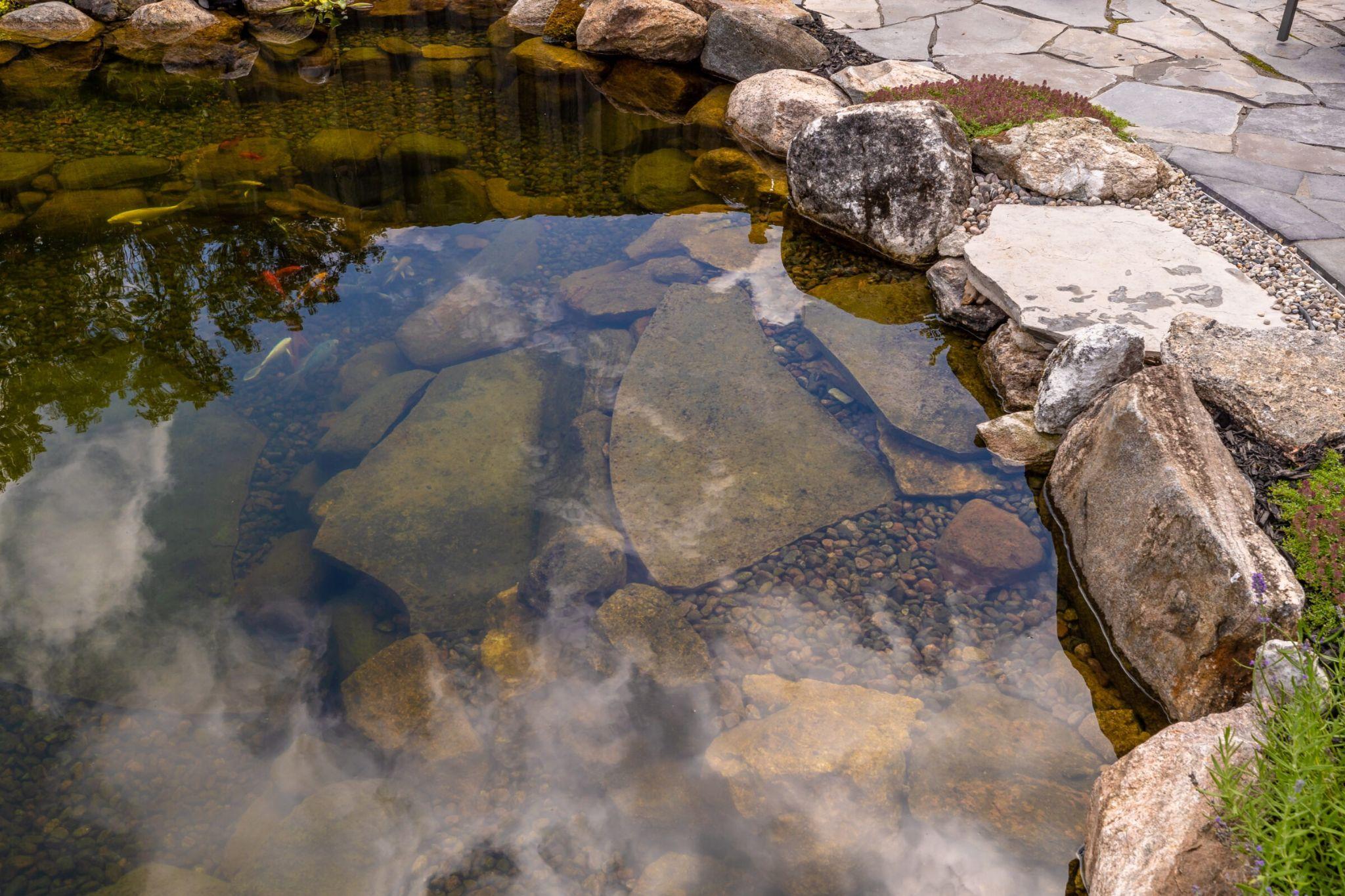 A close view of a clear pond showing large submerged stones, small pebbles, and koi fish near a stone-paved edge.