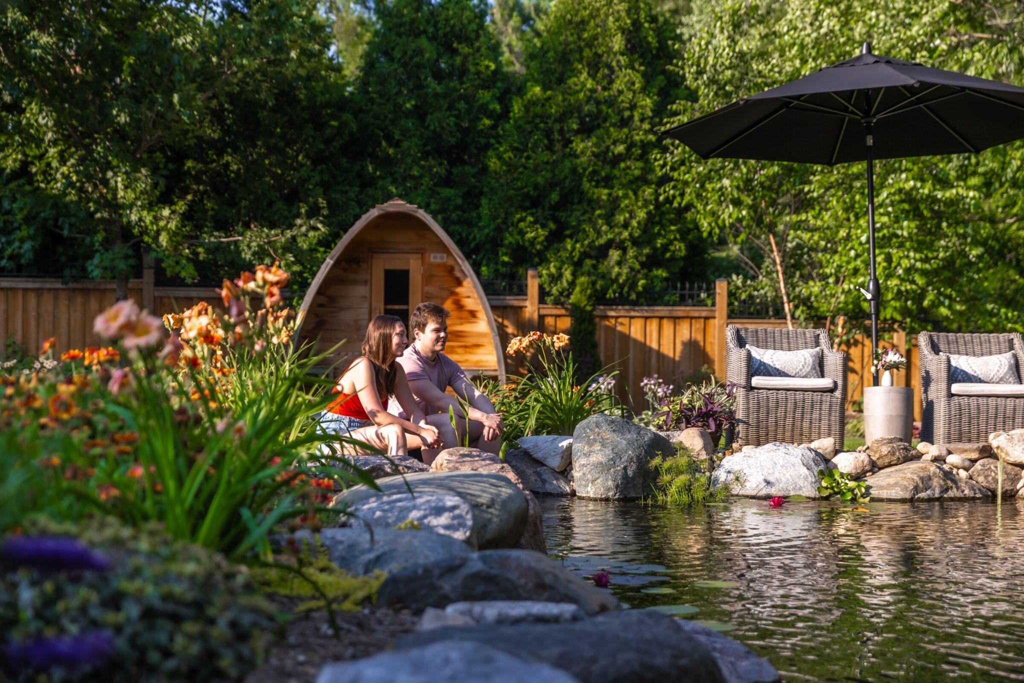 A couple sitting on rocks beside a pond in a landscaped backyard, with flowering plants, outdoor seating, and a wooden sauna structure behind them.