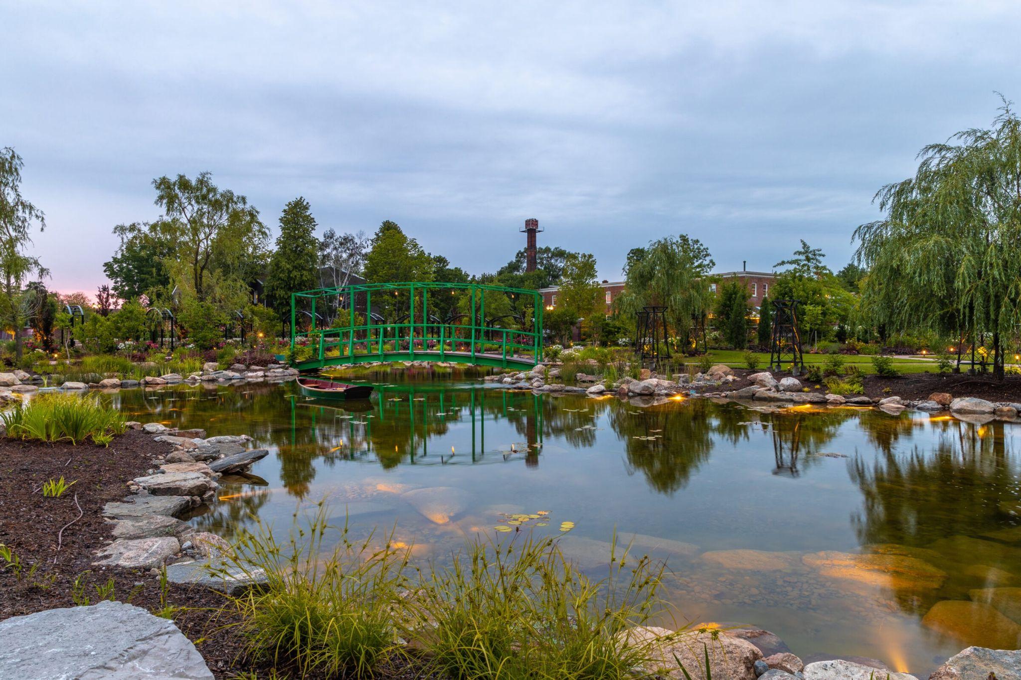A reflective garden pond at dusk with a green metal footbridge, stone edging, and soft landscape lighting.