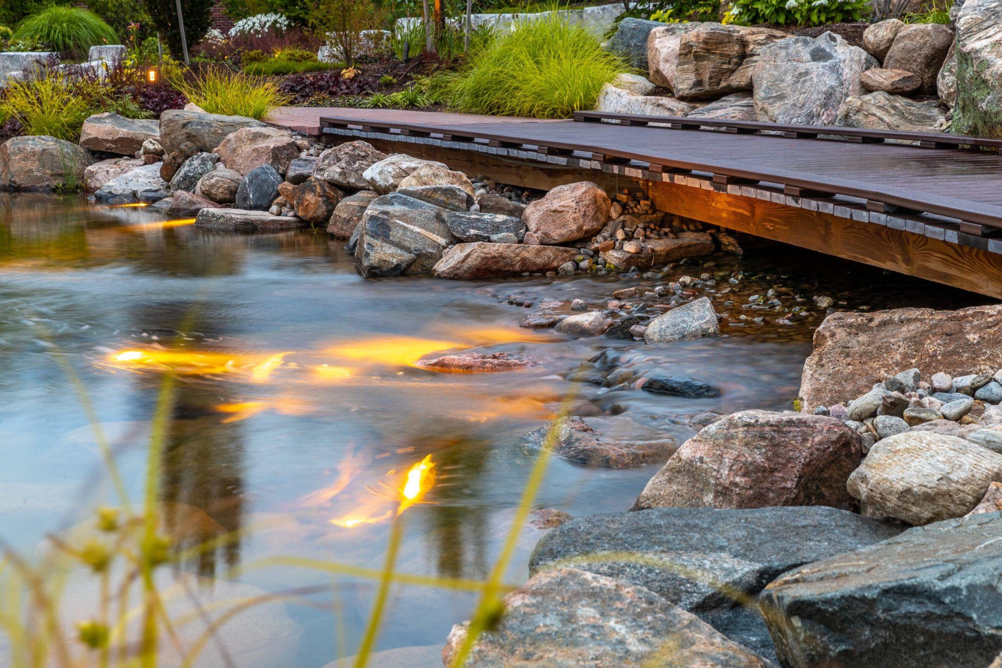 A shallow stream flowing under a wooden footbridge, with warm underwater lights illuminating rocks beneath the moving water.