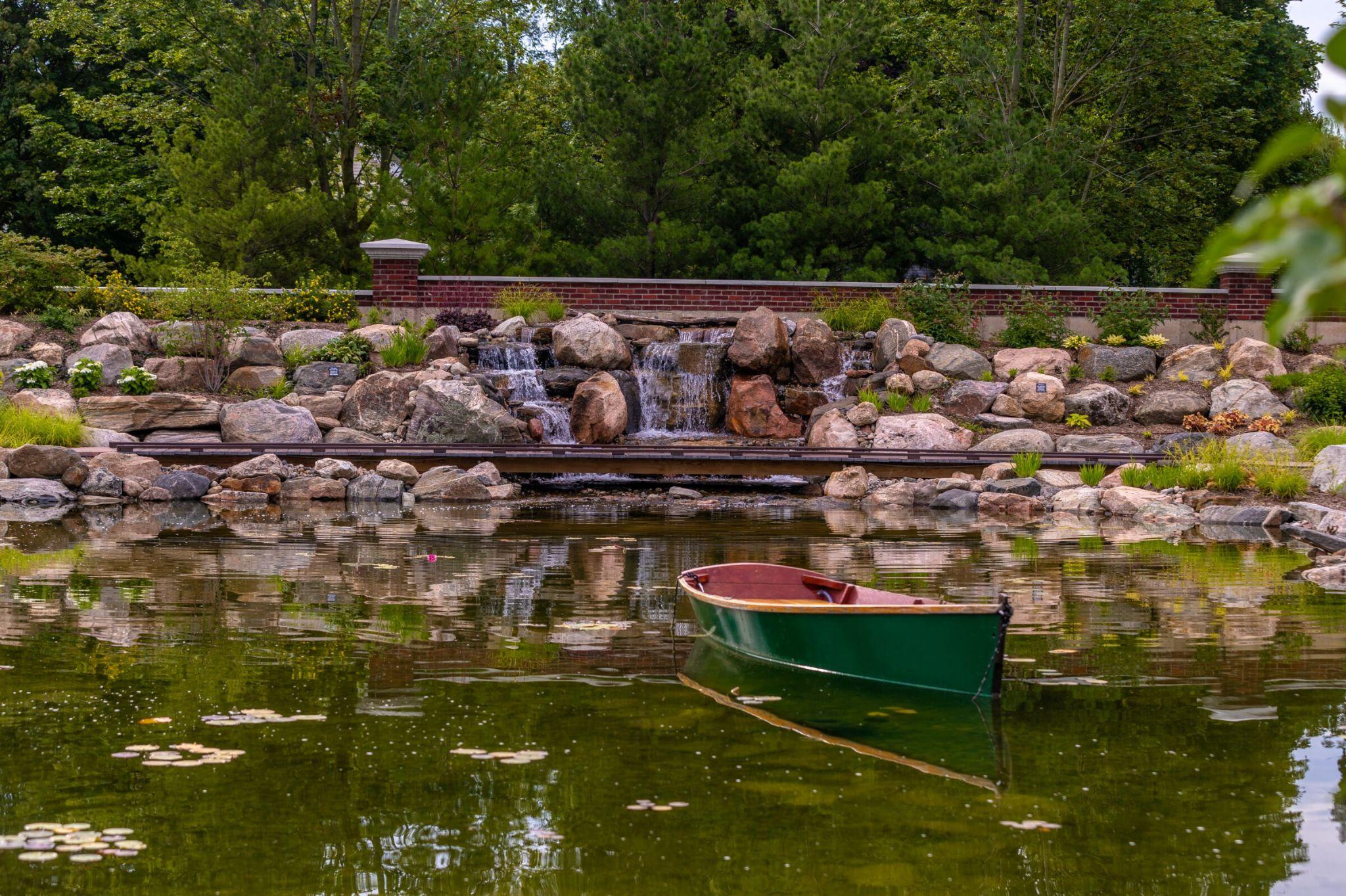 A small green rowboat floating on a pond in front of a stone waterfall, with trees and a brick wall in the background.